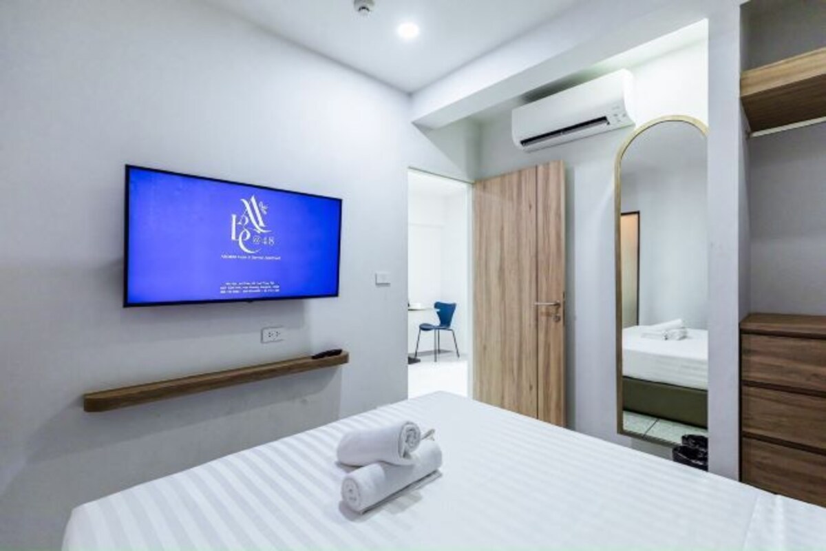 A modern hotel room with a flat-screen TV mounted on the wall, a bed with white linens, a wooden shelf, a mirror, and an air conditioning unit.