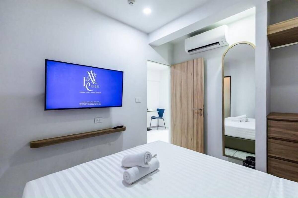 A modern hotel room with a flat-screen TV mounted on the wall, a bed with white linens, a wooden shelf, a mirror, and an air conditioning unit.