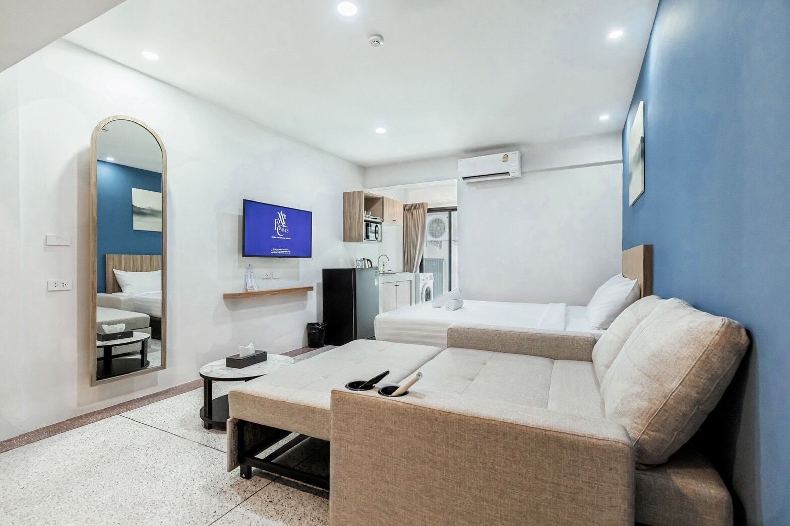 A modern hotel room with a sofa, a TV, a mirror, a small table, and a kitchenette.