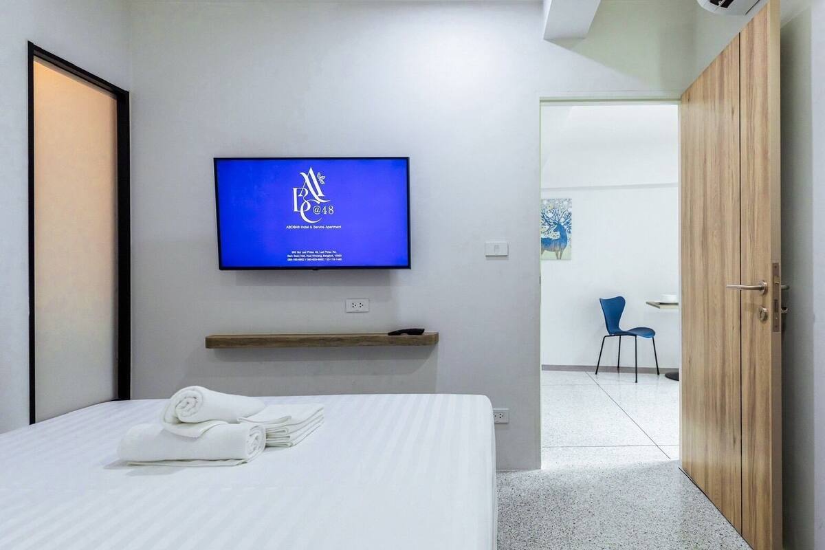 A hotel room with a bed, a TV mounted on the wall, a wooden shelf, and a door leading to another room.