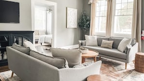 A living room with a grey sofa, a round coffee table, and a fireplace. There is a large window with curtains and a plant on the windowsill.