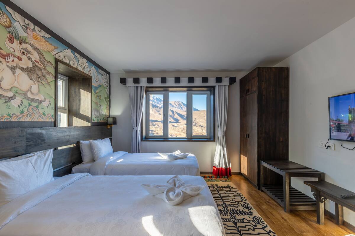 A hotel room with two beds, a large window with a mountain view, a wooden headboard, a TV, and a wooden bench.