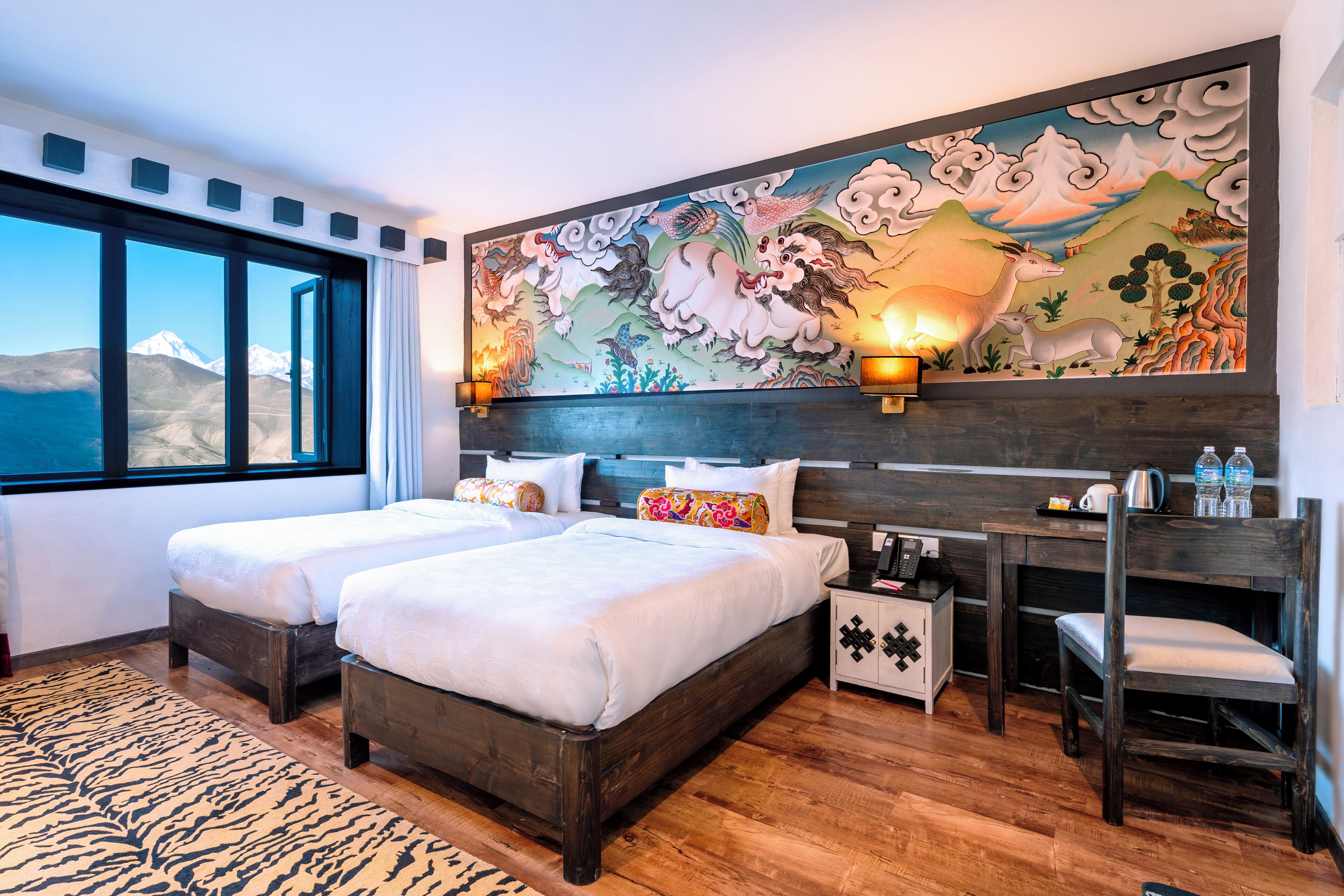 A hotel room with two beds, a desk, a chair, a nightstand, and a large mural on the wall.