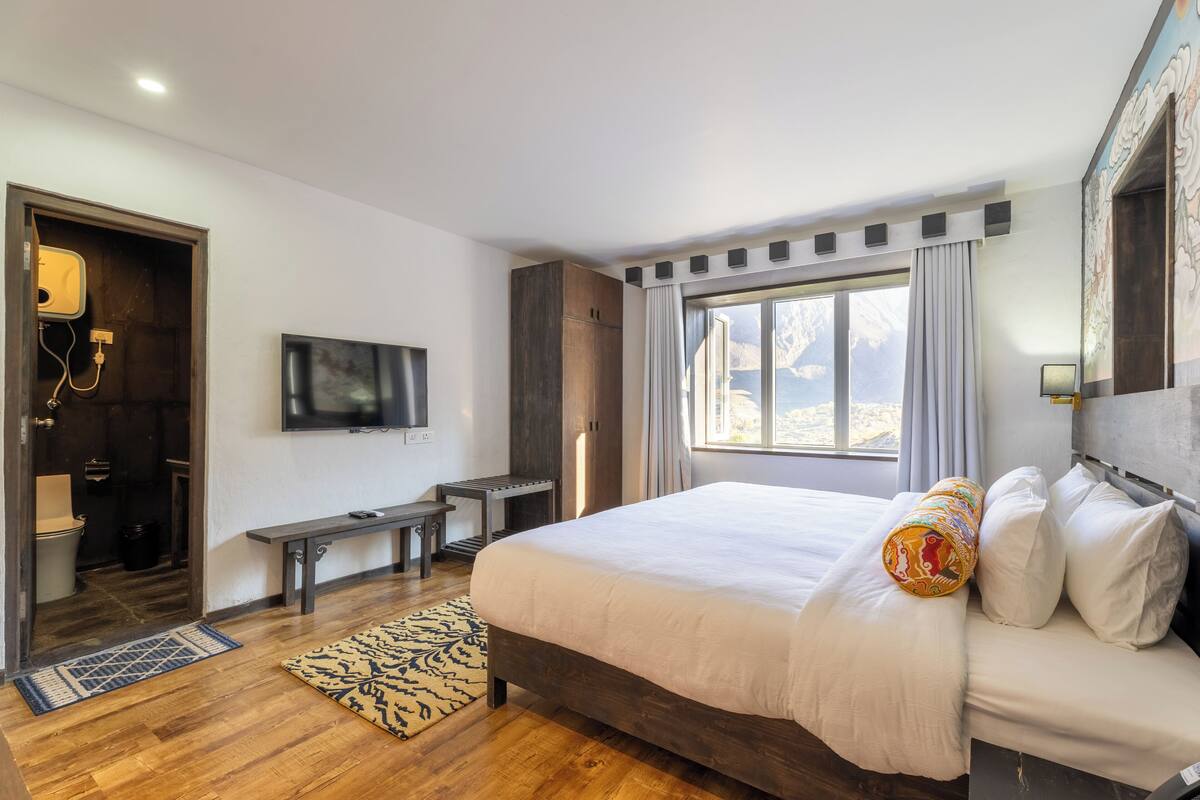 A modern bedroom with a large bed, a TV, a bench, and a view of mountains.