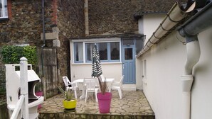 Une petite cour avec un sol en pierre, des chaises blanches, une table et des plantes en pot.