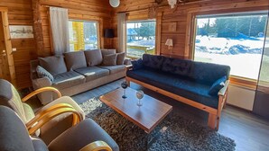 A cozy log cabin interior with a sofa, chairs, a coffee table, and a large window offering a snowy view.