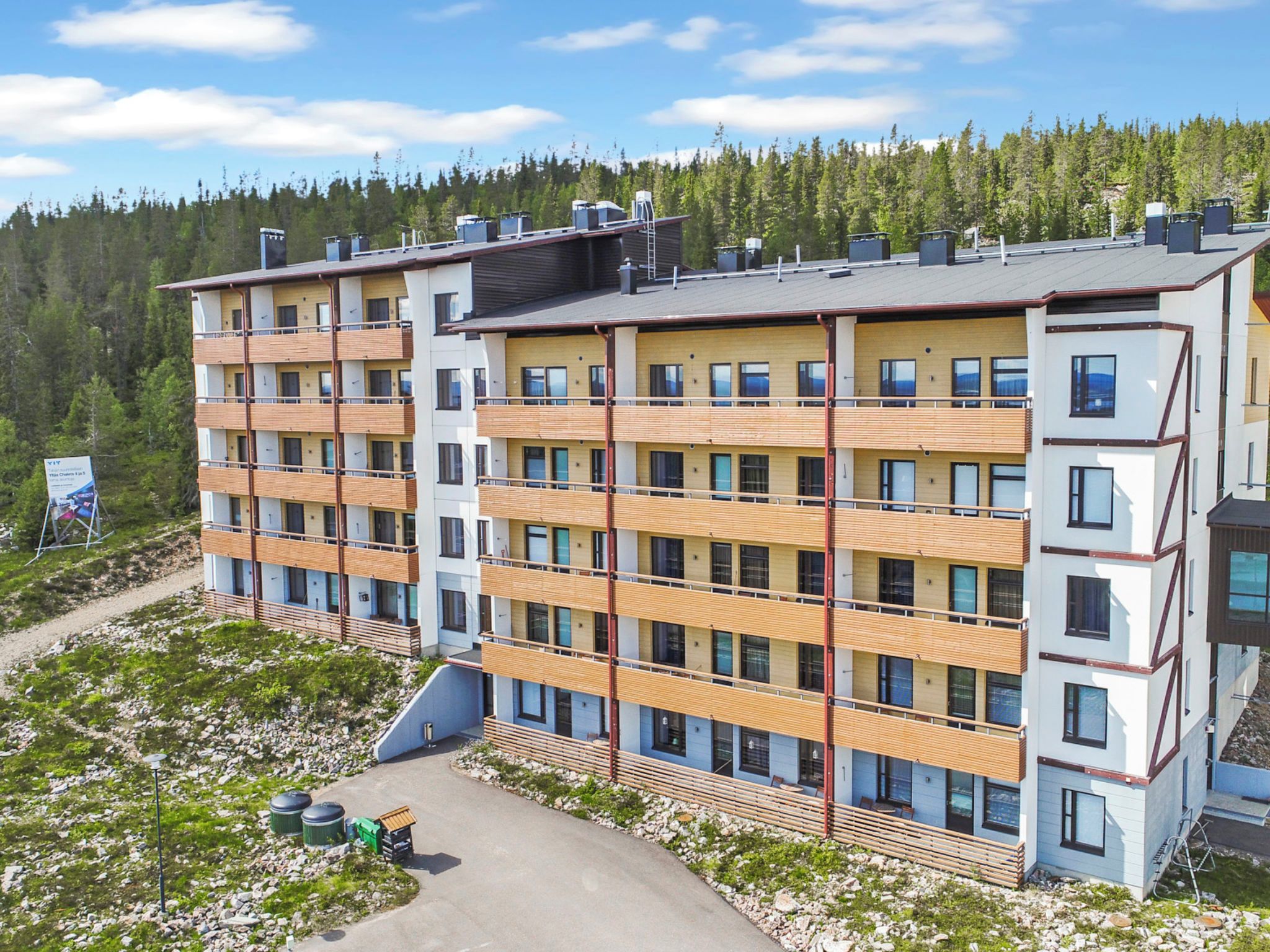 Un edificio a più piani con balconi, un vialetto lastricato e uno sfondo boschivo.