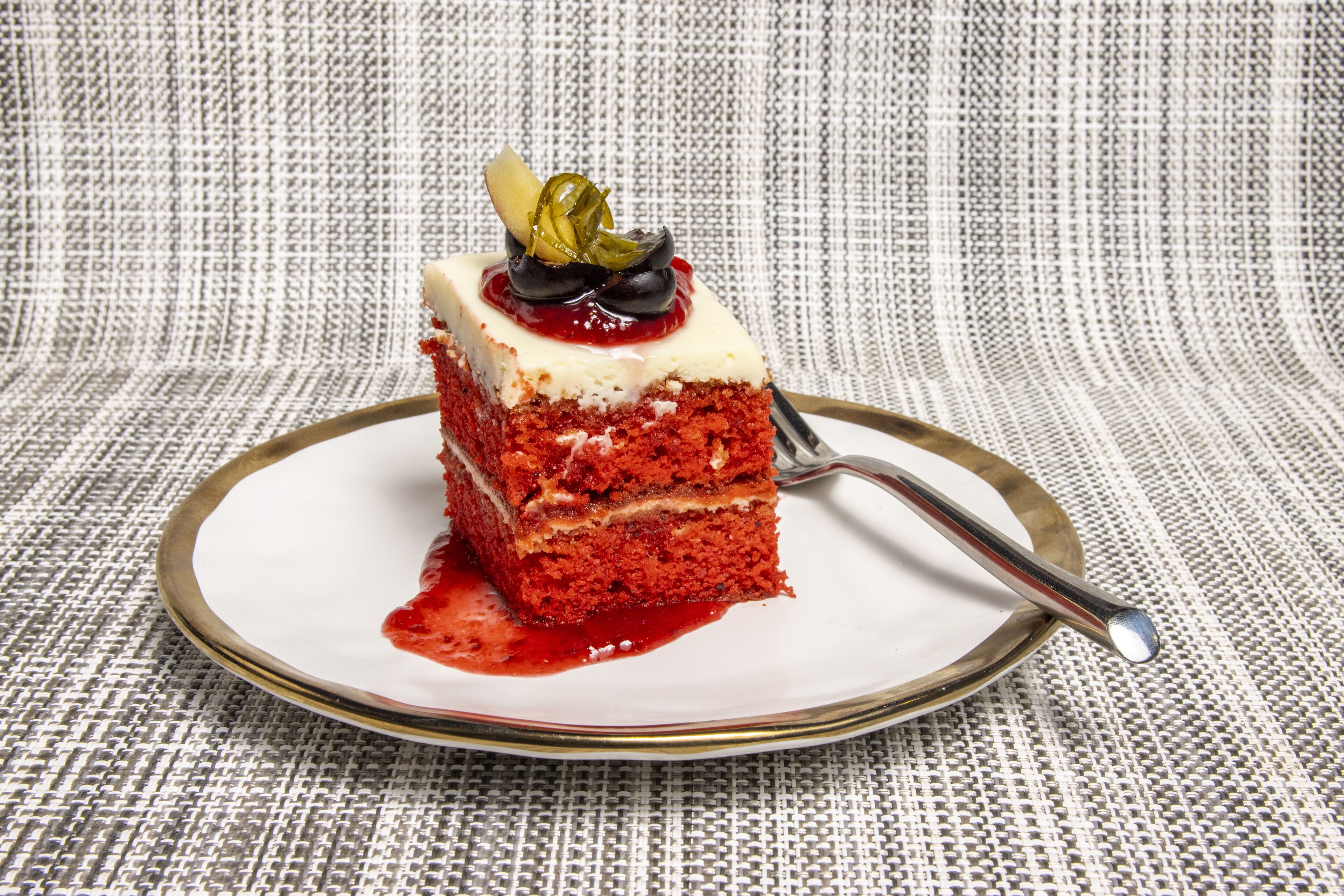 A slice of red velvet cake with white cream and a cherry on a white plate with a gold rim, placed on a textured fabric surface.