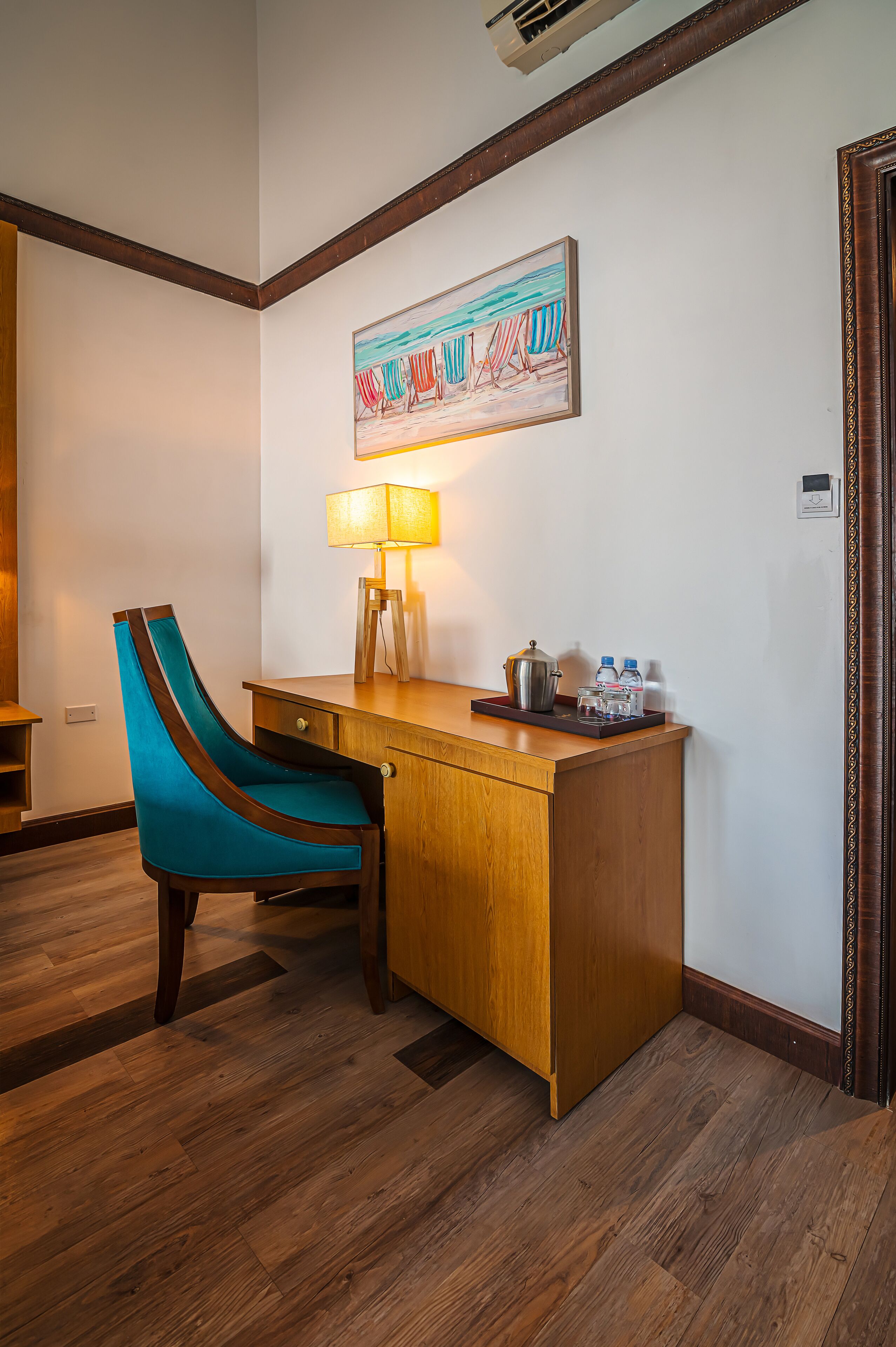 A hotel room with a wooden desk, teal upholstered chair, a painting on the wall, a lamp, and a water dispenser.