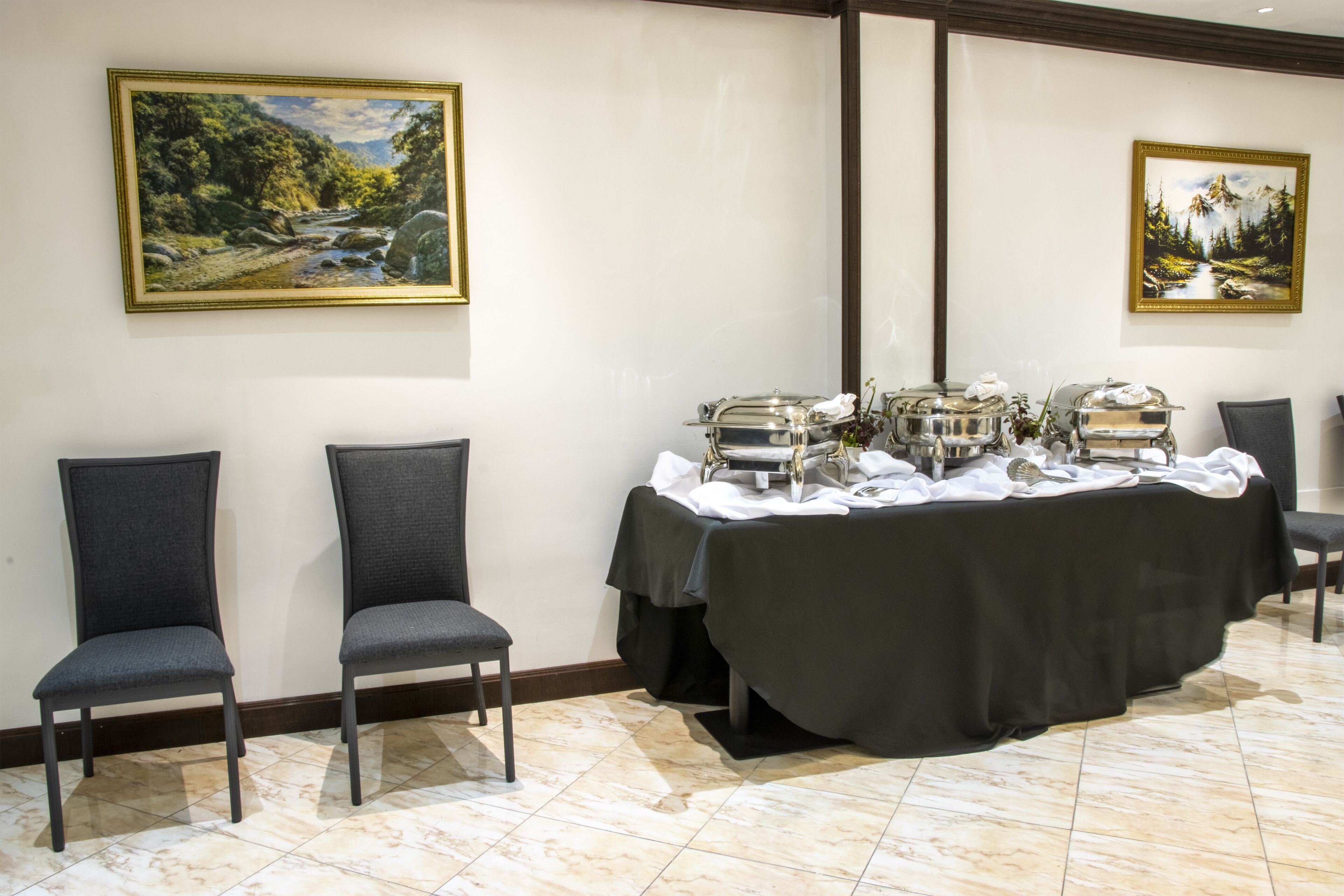 A room with a buffet table, two chairs, and two framed landscape paintings on the walls.