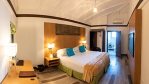 A hotel room with a large bed, bedside lamps, a wooden headboard, a desk with a lamp, a small table, and a door leading to a bathroom. - Sago Palm Hotel (Ocho Rios)