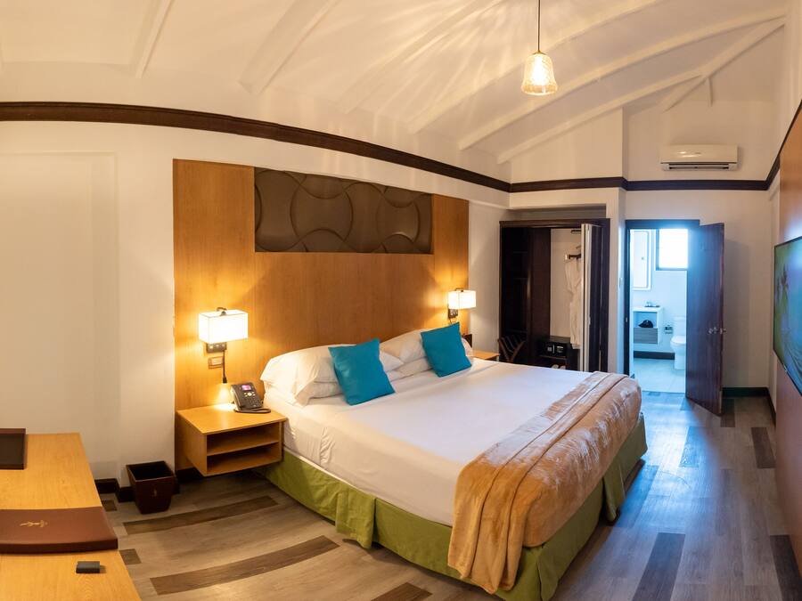 Et hotelværelse med en stor seng, sengeborde med lamper, et træhovedgærde, et skrivebord med lampe, et lille bord og en dør til et badeværelse.