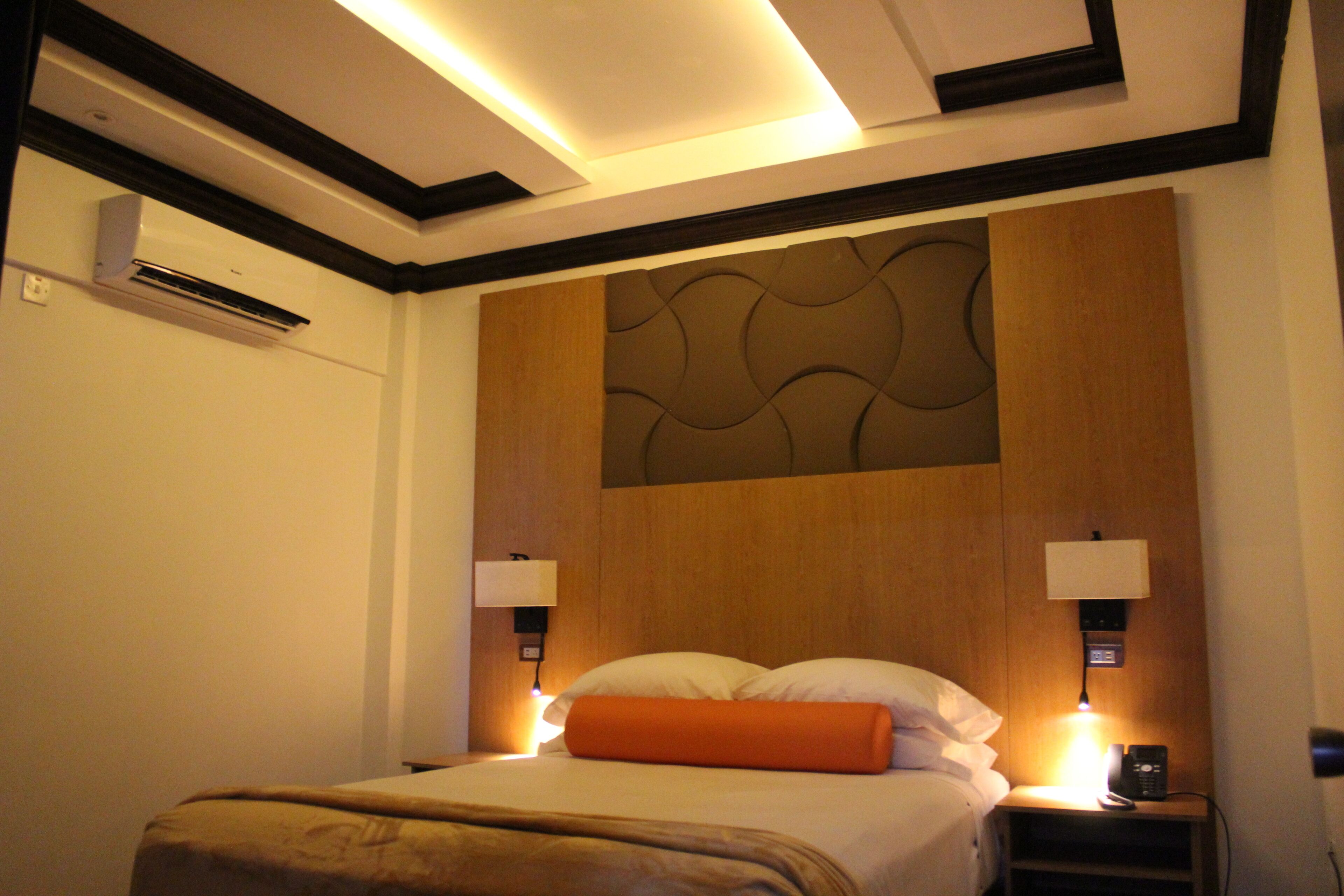 A hotel room with a bed, bedside lamps, an air conditioning unit, and a wall-mounted panel.
