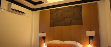A hotel room with a bed, bedside lamps, an air conditioning unit, and a wall-mounted panel.