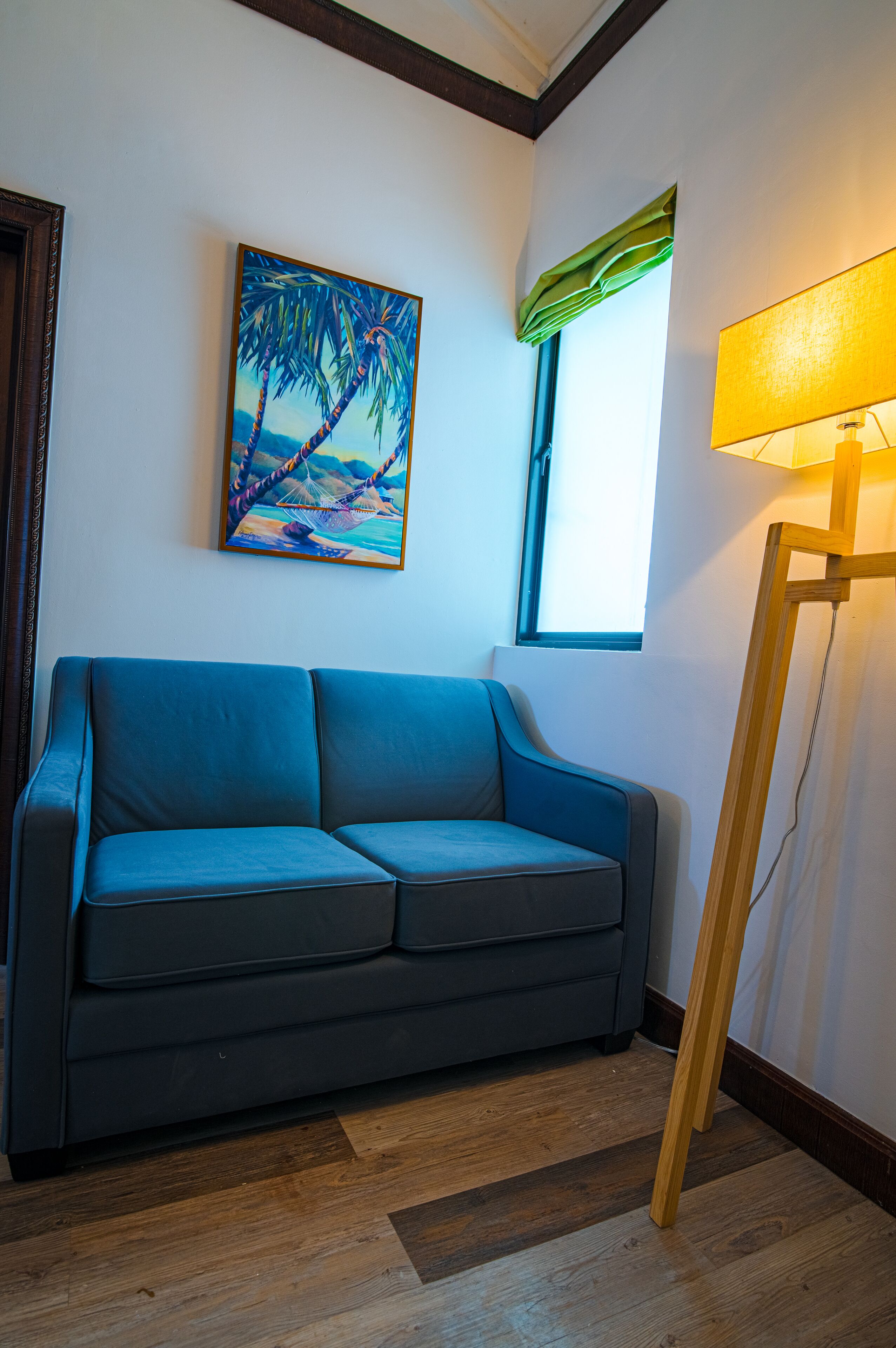 A room with a blue sofa, a painting of a beach scene, a window with a green curtain, and a floor lamp.
