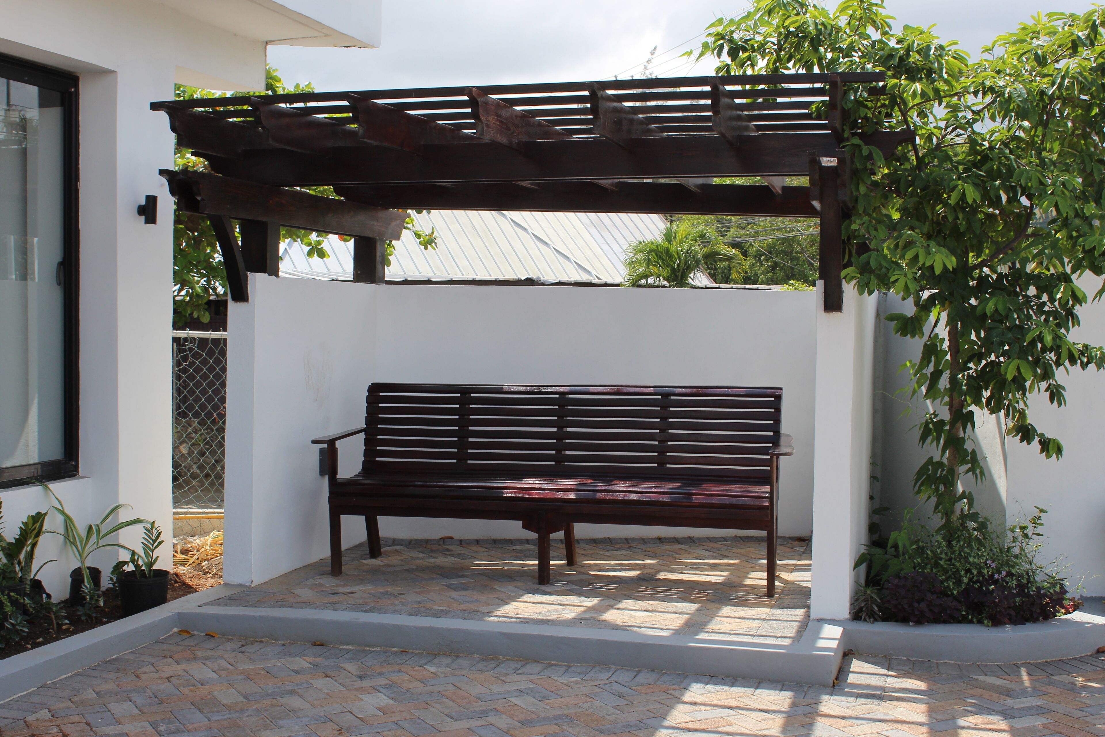 A wooden bench under a pergola in a courtyard with a tiled floor and potted plants.