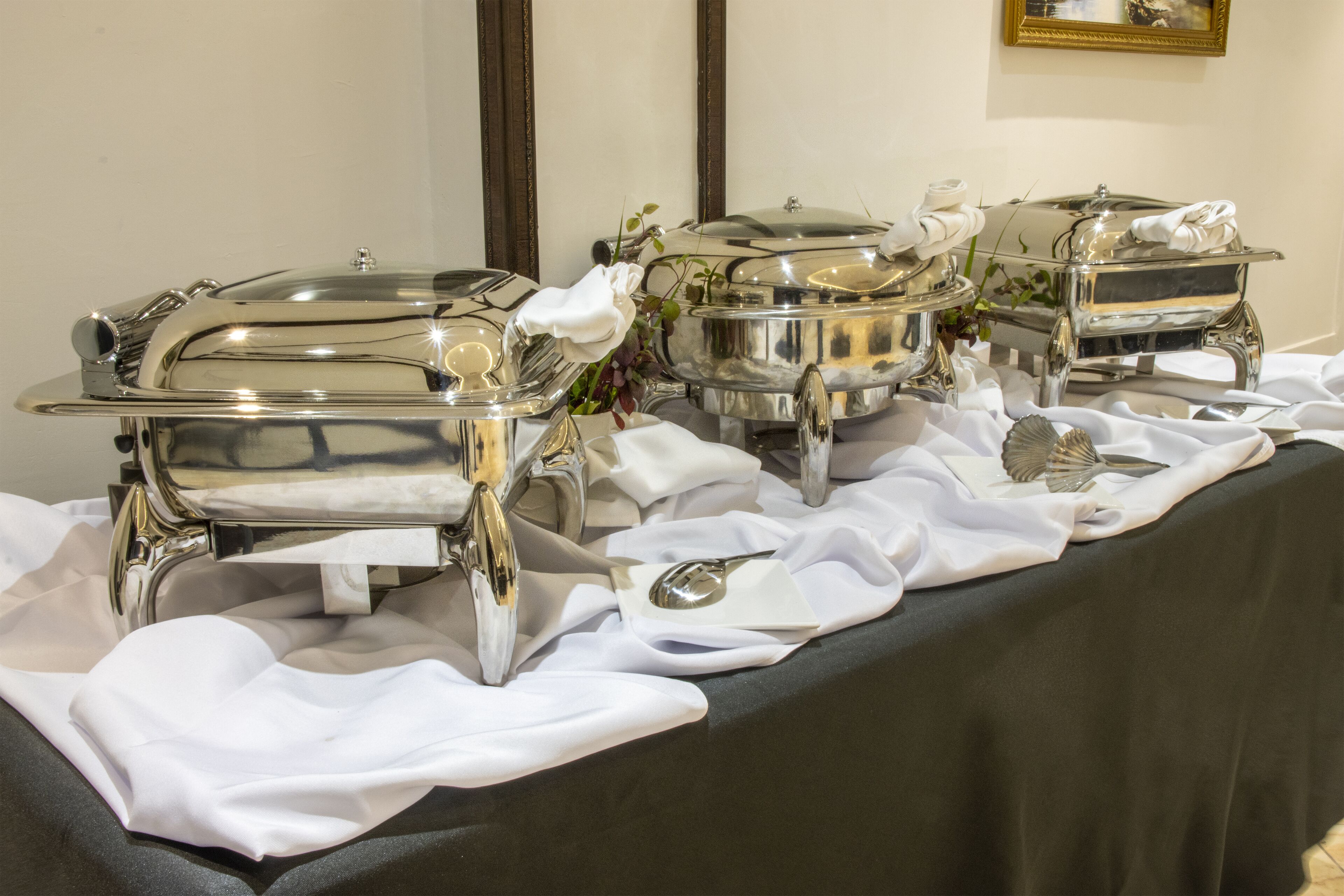 A buffet setup with multiple silver chafing dishes on a table covered with a black tablecloth and white tablecloths.