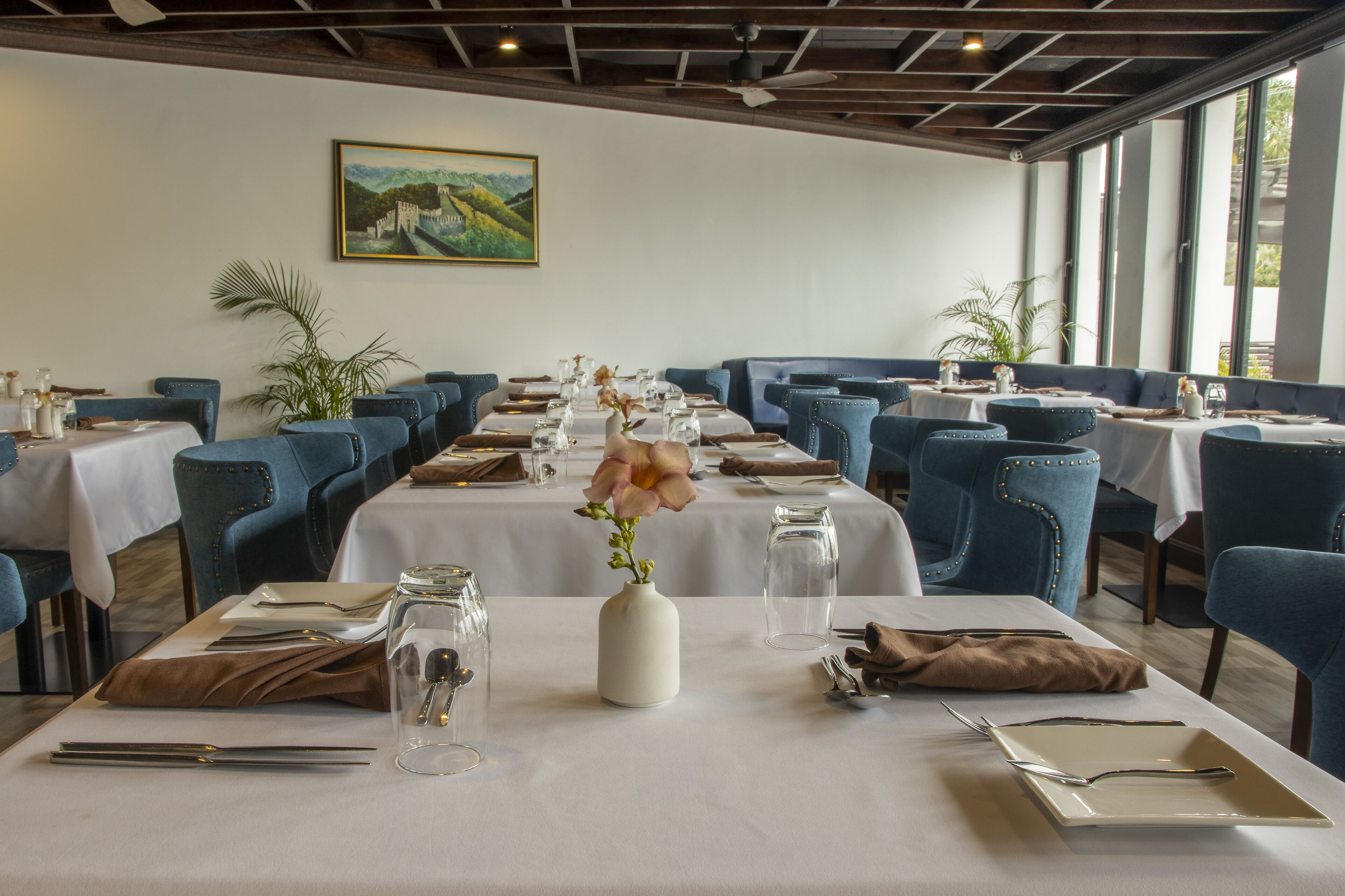 A dining area with tables set for service, featuring blue upholstered chairs, white tablecloths, and a vase with flowers.