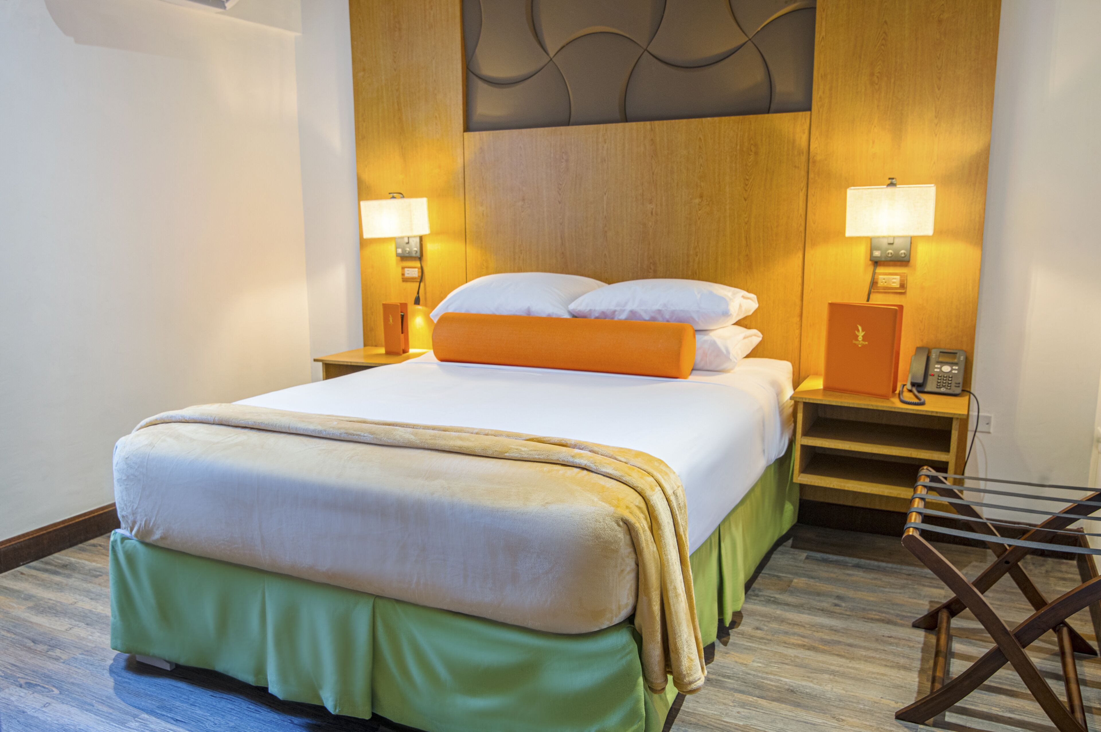 A neatly made bed with a green skirt, two bedside lamps, a telephone, and a wooden nightstand.