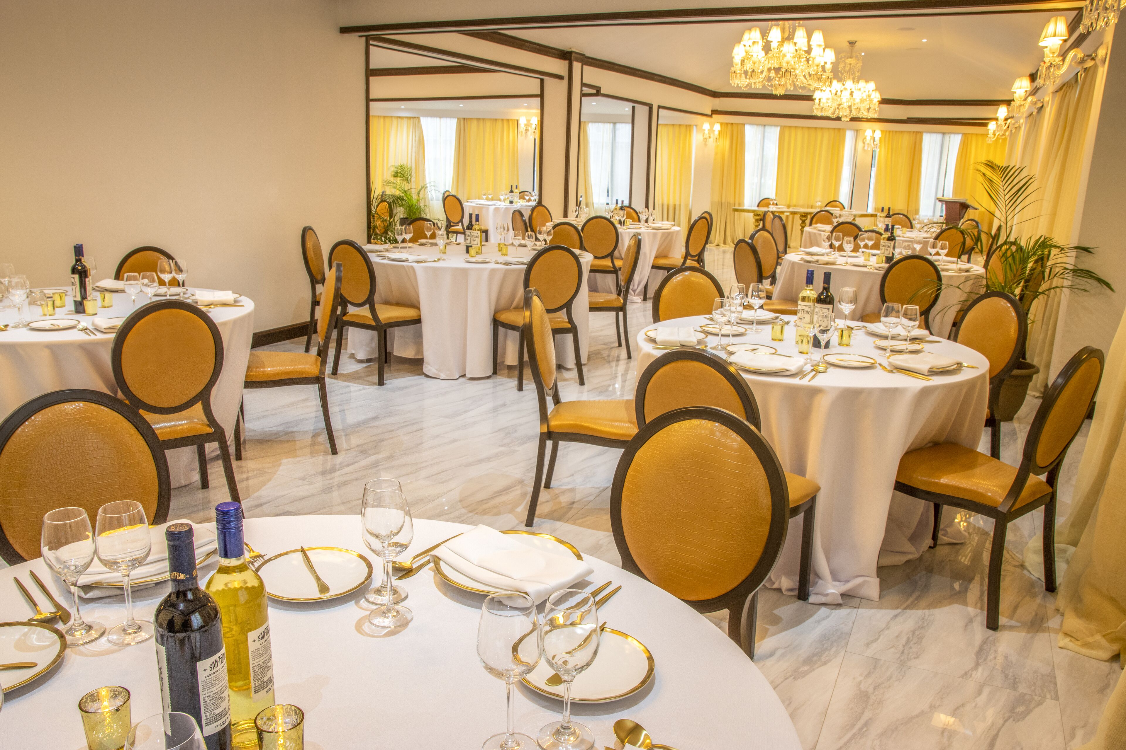 Un comedor de hotel con varias mesas redondas dispuestas para comer, cada una con mantel blanco, platos con borde dorado y sillas con cojines dorados.