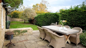 Une terrasse avec une table et des chaises en osier, entourée de verdure et d’un mur en pierre.