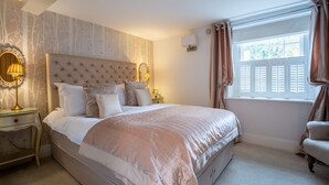 Une chambre à coucher avec une tête de lit capitonnée, un grand lit avec de nombreux oreillers, des lampes de chevet, une fenêtre avec des stores et une chaise près de la fenêtre.