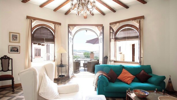 Un salon spacieux avec un canapé de couleur turquoise, des fauteuils blancs et une table basse en bois. Il y a deux grandes fenêtres avec des volets en bois, un lustre et une vue sur l’extérieur.