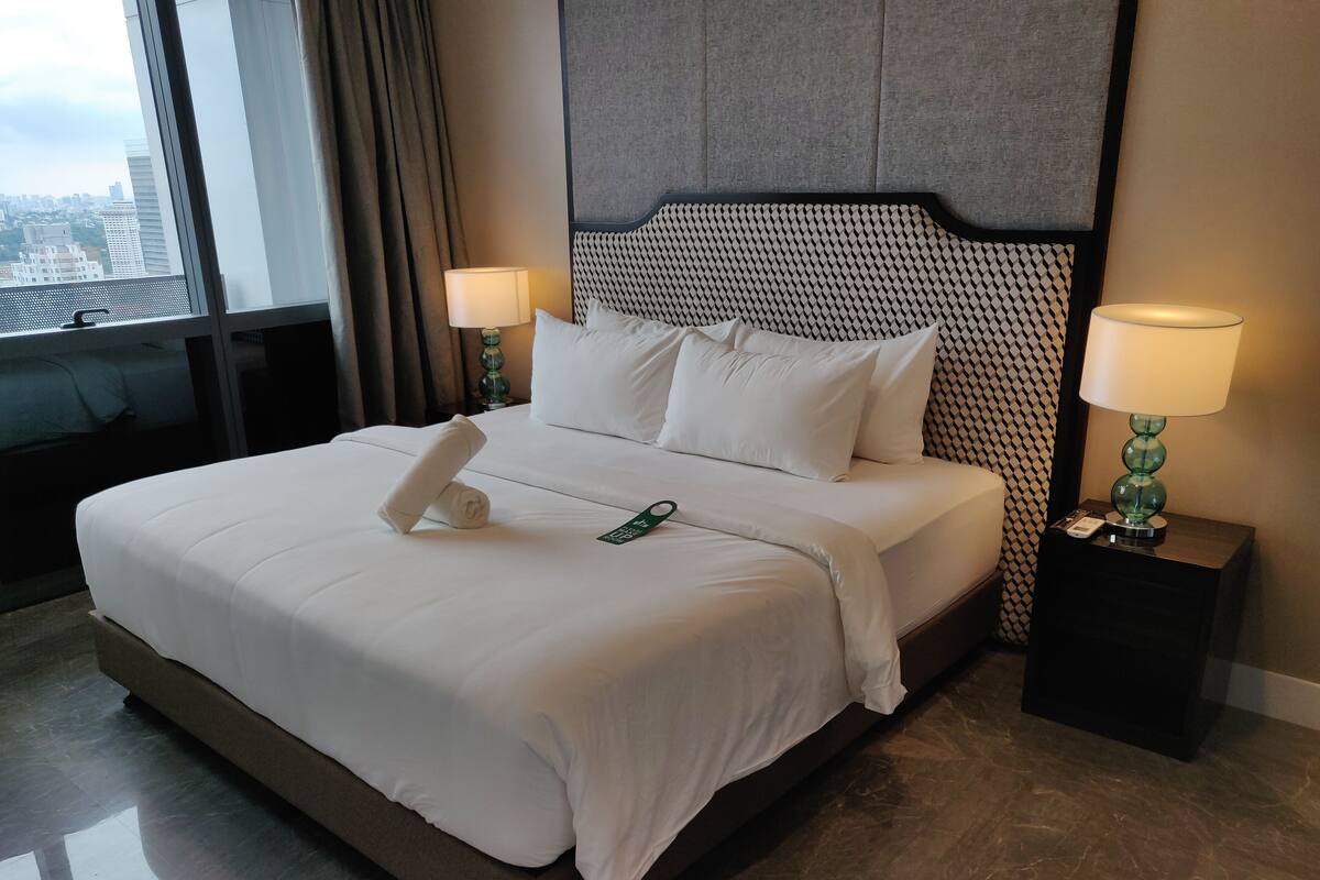 A neatly made bed with white linens, two bedside lamps, and a view of the city through a large window.