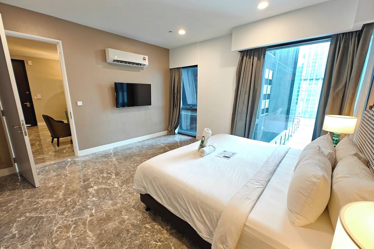 A hotel room with a large bed, a flat-screen TV mounted on the wall, an air conditioning unit, a window with curtains, and a view of the cityscape.