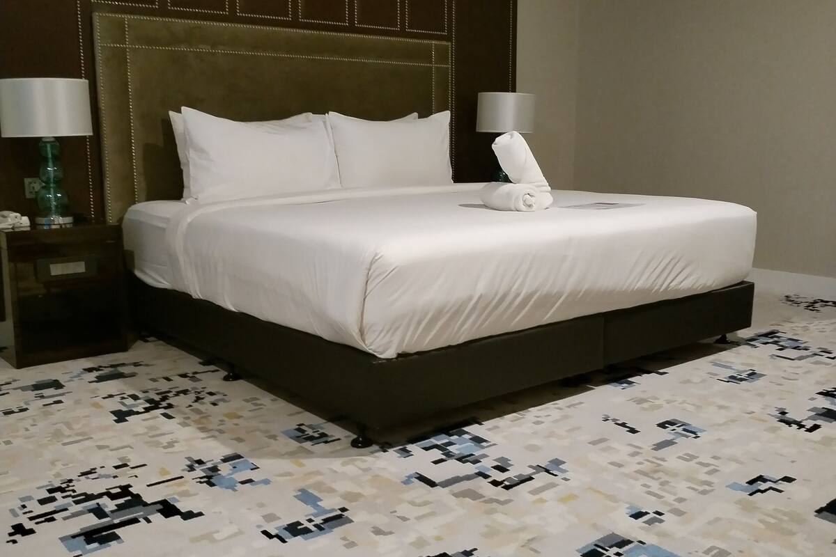 A hotel room with a large bed, a headboard with a geometric pattern, a bedside table with a lamp, and a patterned carpet.