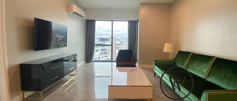 Una sala de estar moderna con un televisor de pantalla plana, un sofá verde, una mesa de centro y vistas de la ciudad a través de ventanas amplias.