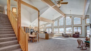 Een ruime woonkamer met een hoge plafond, grote ramen, een houten trap en een centraal eiland met zitplaatsen.