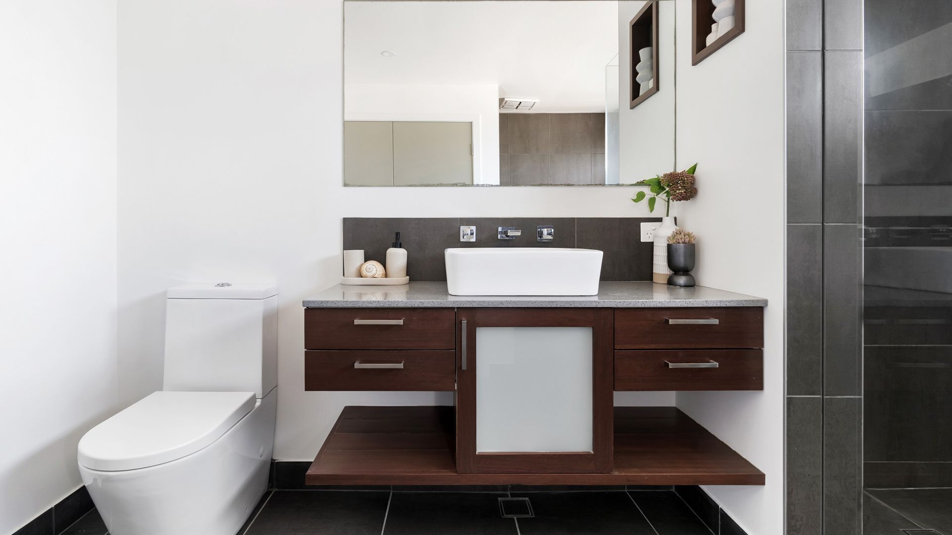 Un baño moderno con inodoro blanco, tocador de madera oscura y lavamanos blanco, y un espejo sobre el tocador.
