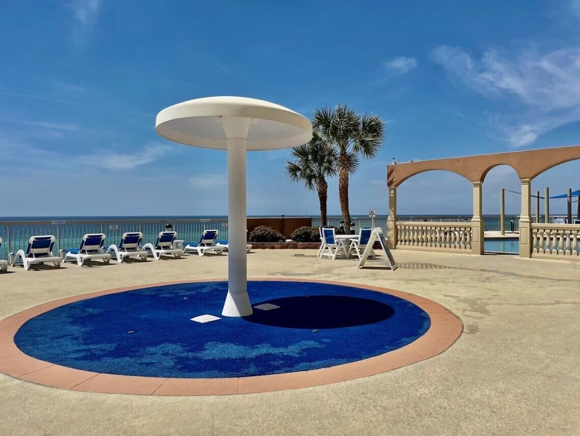 A pool area with a central fountain, lounge chairs, and a pergola with a view of the sea.