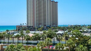 Un hotel alto con varios balcones, cerca de una playa con palmeras y coches aparcados.