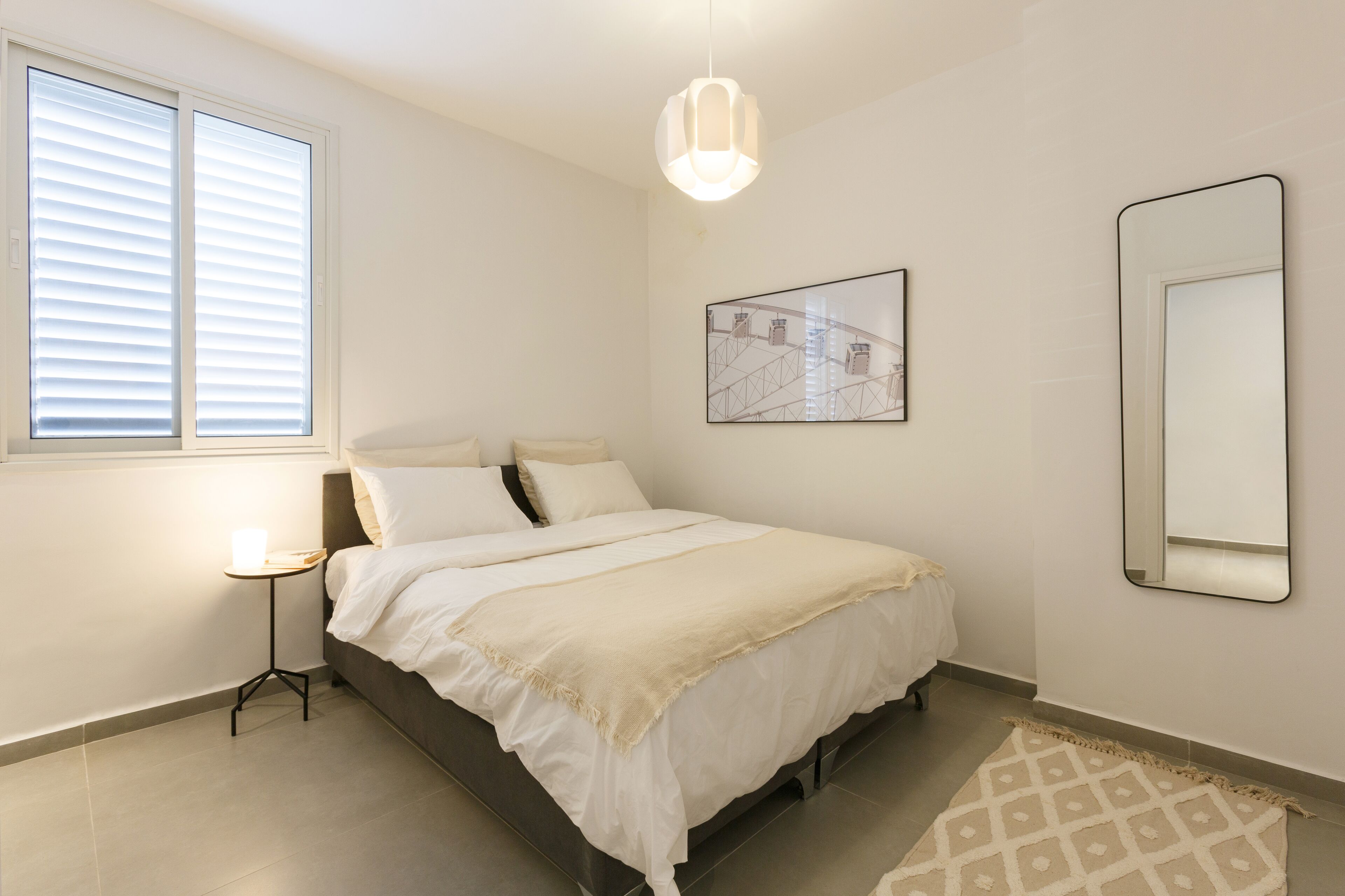 A bedroom with a bed, a bedside table with a lamp, a framed picture on the wall, and a mirror.