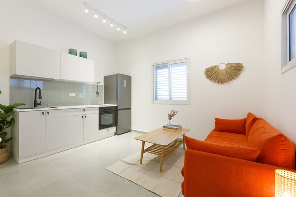 A modern kitchen with white cabinets, a built-in oven, and a refrigerator. There is a wooden table with a vase and books, and an orange sofa in the living area.