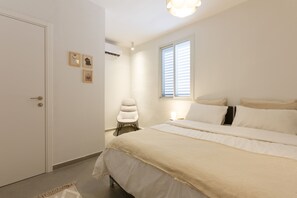 A bedroom with a bed, a chair, a window with blinds, and a door. - AirTLV - Kamuel Apartments (Tel Aviv)