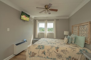 Un dormitorio con cama, ventilador de techo, ventana con cortinas, televisor montado en la pared y unidad de aire acondicionado.