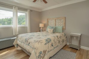 A bedroom with a bed, a nightstand, a lamp, a window with curtains, and a view of greenery outside.