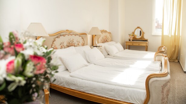 Una cama doble con estructura de madera y ropa de cama blanca, una mesita de noche con espejo, y una ventana con cortinas.