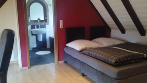Une petite chambre mansardée, chaleureuse et accueillante, avec un lit, une chaise et une salle de bain visible à travers une porte ouverte.