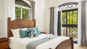 Un lit bien fait, avec une tête de lit en bois, une literie blanche et des coussins décoratifs. Il y a deux tables de chevet avec des tiroirs en bois, et une paire de serviettes enroulées est posée sur le lit. La pièce est dotée de grandes fenêtres avec des rideaux, et offre une vue sur la verdure à l’extérieur.