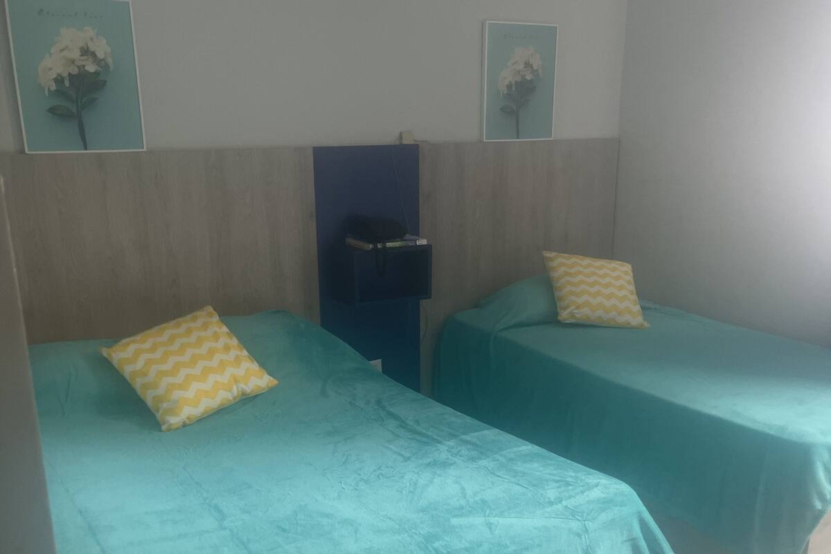 A room with two beds, each with a teal bedspread and a yellow pillow. There is a blue cabinet against the wall and a framed picture above it.