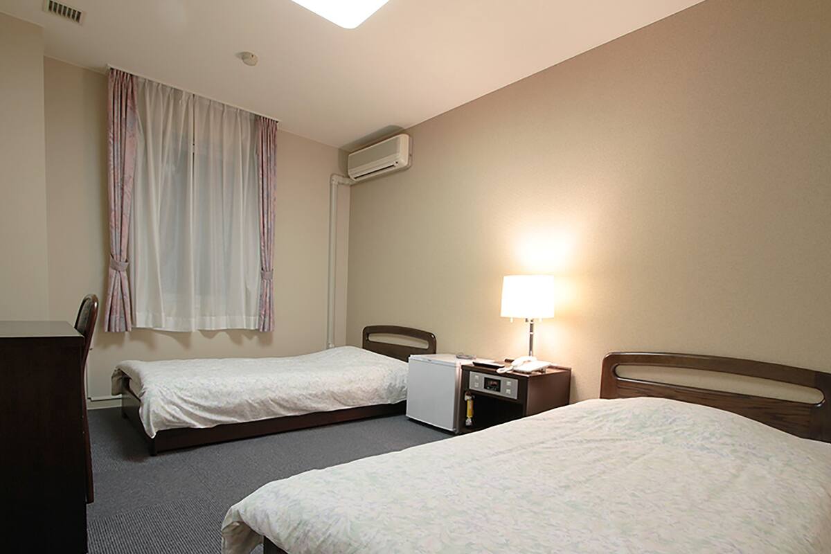 Een hotelkamer met twee eenpersoonsbedden, een bureau, een stoel, een airconditioning, een lamp en een raam met gordijnen.