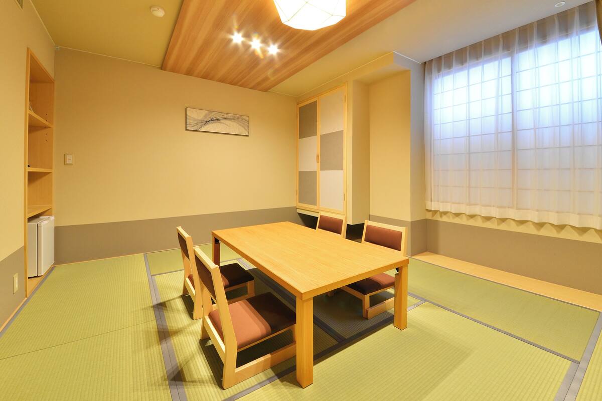 Een kamer met een houten tafel, stoelen, tatami vloerbedekking, een plafondlamp, een raam met gordijnen en een aan de muur bevestigd kunstwerk.