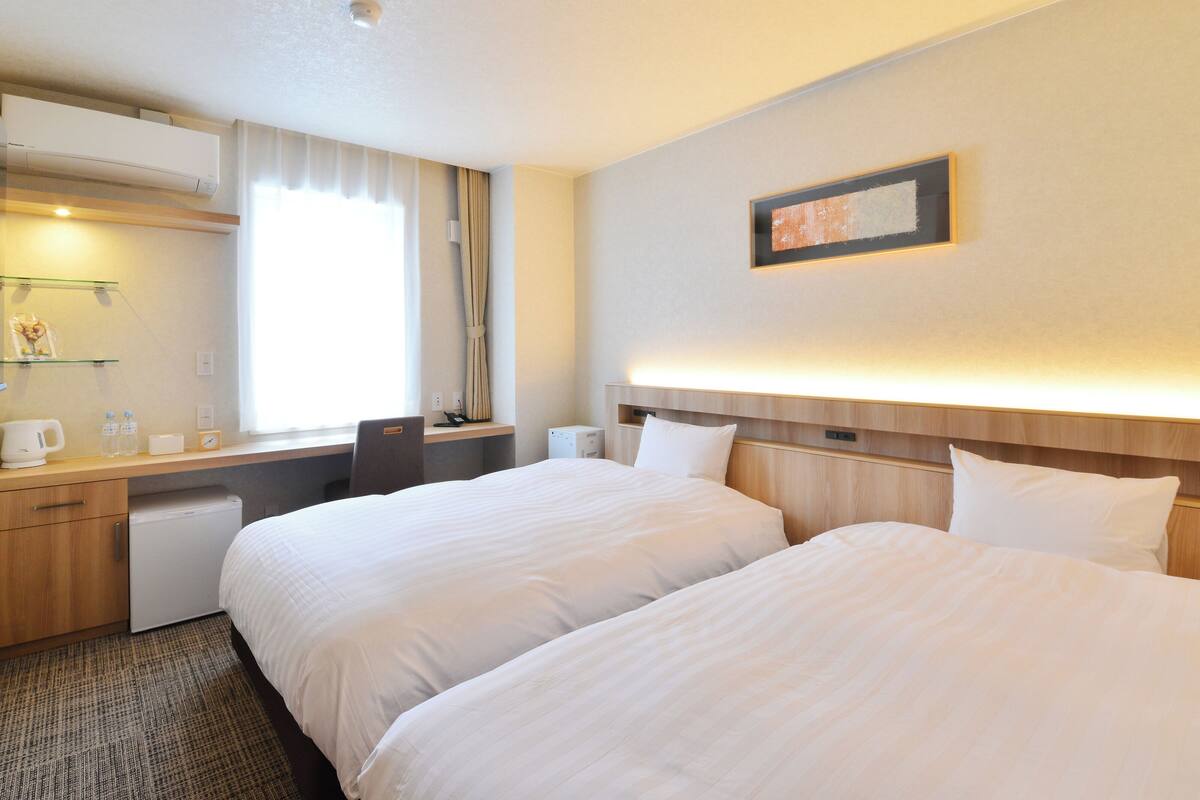 Een hotelkamer met twee bedden, een bureau, een stoel, een klein tafeltje, een spiegel en een raam met gordijnen.