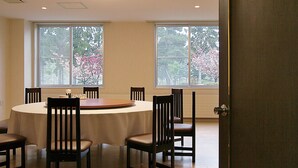 A dining area with a round table and six chairs, large windows with a view of trees, and a door to the right.
