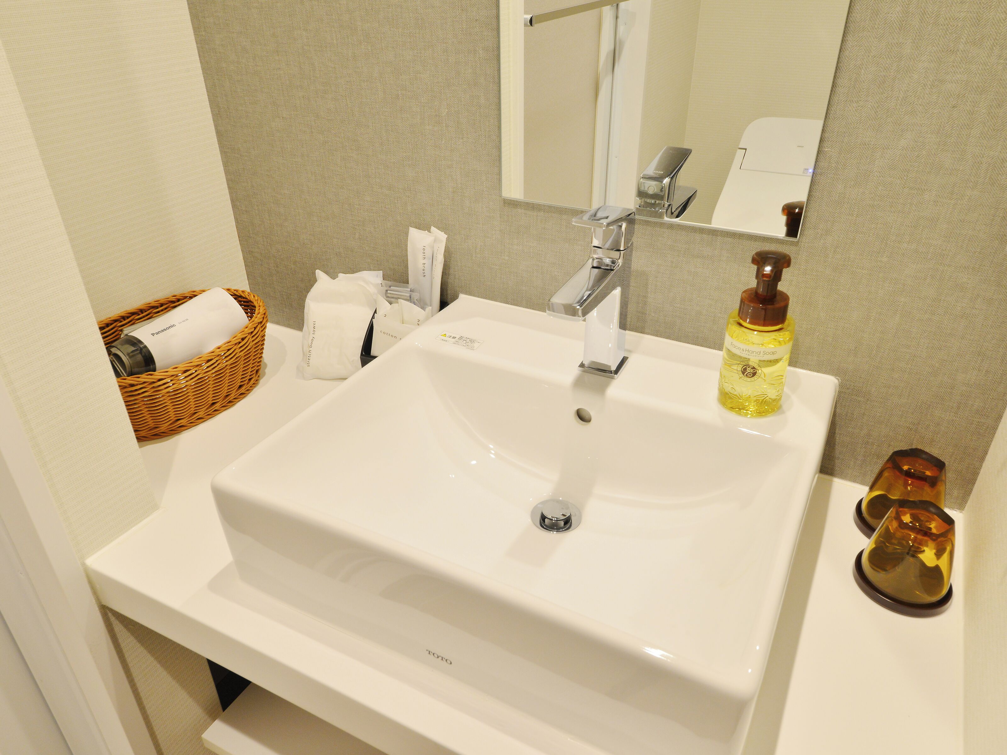 A bathroom with a white sink, a mirror, a towel dispenser, a soap dispenser, and a wicker basket with a towel.