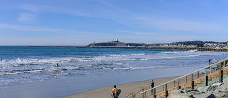 A beach with a wooden staircase leading down to the shore, a rocky outcrop in the foreground, and a distant view of a lighthouse and buildings.